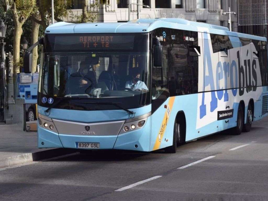 一辆蓝白相间的航空巴士停靠在巴塞罗那西班牙广场，周围是乘客，喷泉雄伟壮观，背景是蒙朱伊克山。