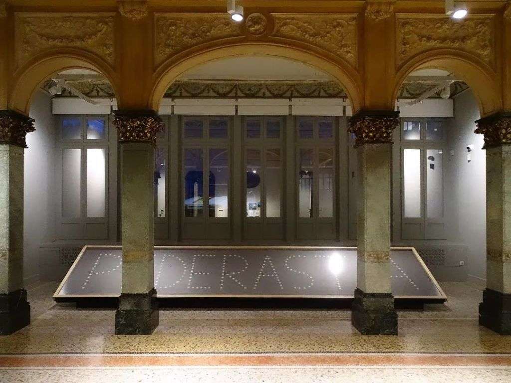 阿贝尔-阿兹科纳（Abel Azcona）名为 '阿门 '的表演图片，展示了艺术家在展览空间中挑战宗教或社会规范的挑衅姿态，以及可能的象征元素，如文本、宗教物品或他自己的身体。.