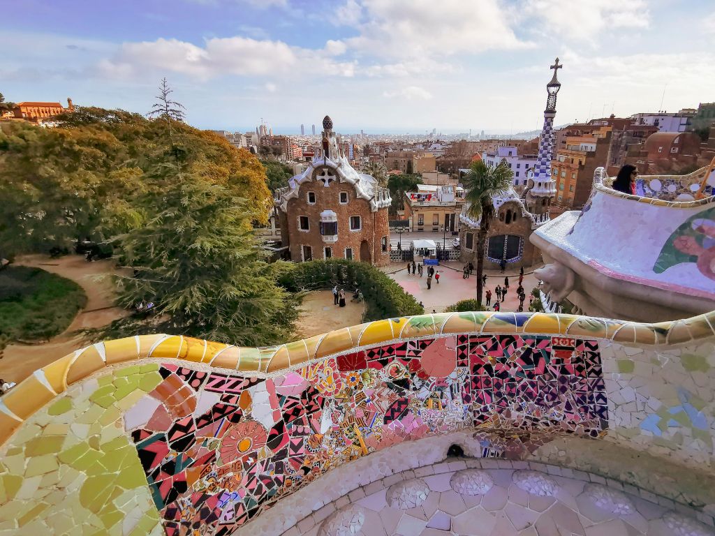 Vista panorámica de Barcelona desde el Parc Güell, mostrando la ciudad extendiéndose hacia el mar bajo el cielo claro, con elementos de la arquitectura de Gaudí en primer plano.