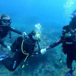En las profundidades de las aguas del montgrí, dos instructores de buceo enseñan a una chica a bucear aguantándole la mano y la bombona para que apreanda a posicionarse estando sumergida.