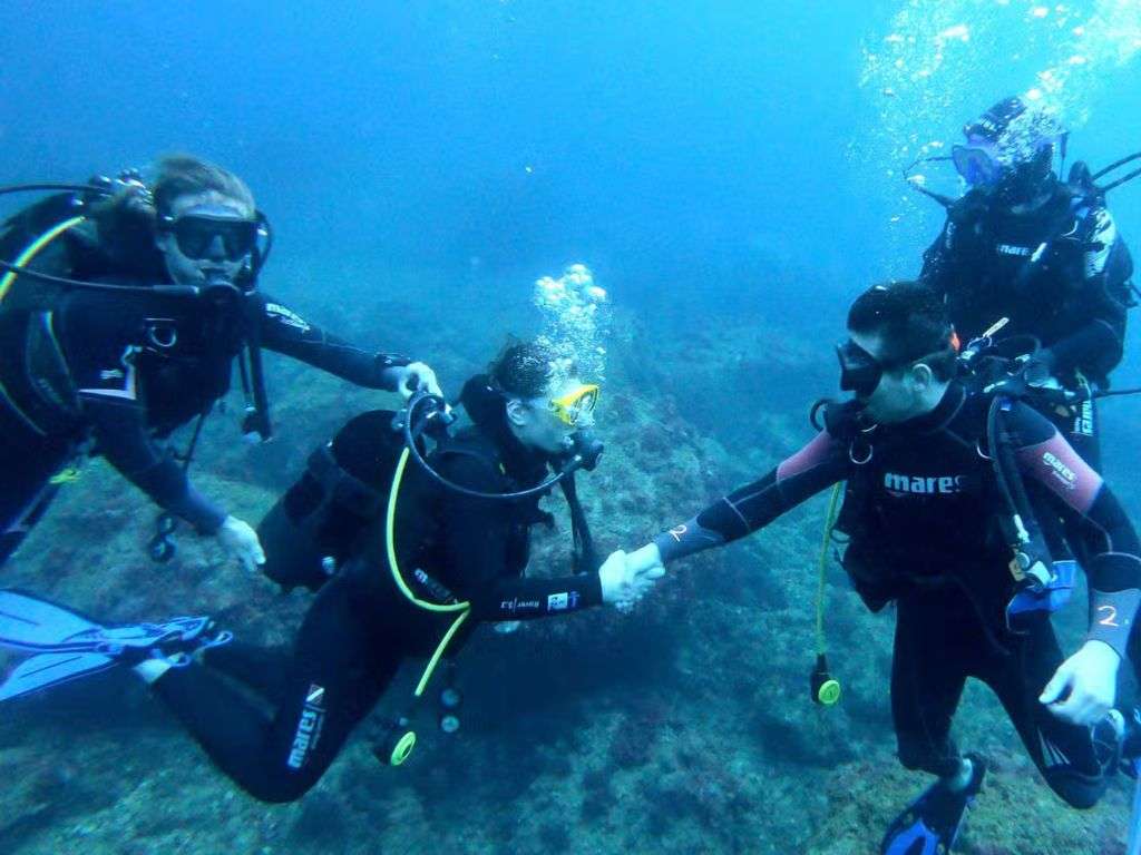 En las profundidades de las aguas del montgrí, dos instructores de buceo enseñan a una chica a bucear aguantándole la mano y la bombona para que apreanda a posicionarse estando sumergida.